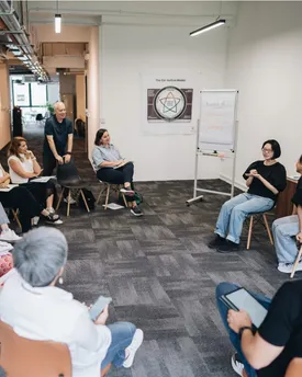 A workshop group seated in a circle in an open-plan office, beside a flip chart reading “Rule #1: nobody gets to be wrong”.