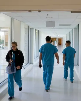 Three healthcare workers in teal scrubs walking through a bright, glass-walled hospital corridor.