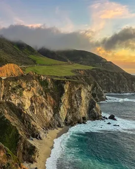The Big Sur coastline at sunset, green headland above steep cliffs with surf breaking on the beach below.