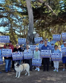 Campaign volunteers and a dog gathered under a cypress tree, holding “Mike Moeller for Mayor — Moving Marina Forward, Together” signs.