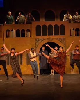 Dancers mid-leap in period costume on a theatre stage, in front of a two-storey set of arched windows and balconies.