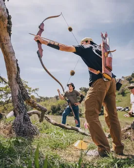 An archer at full draw with a traditional recurve bow in an oak meadow, two others watching from behind.